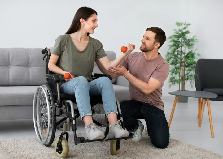 A support worker kneeling beside a young woman in a wheelchair as she lifts a hand weight at home