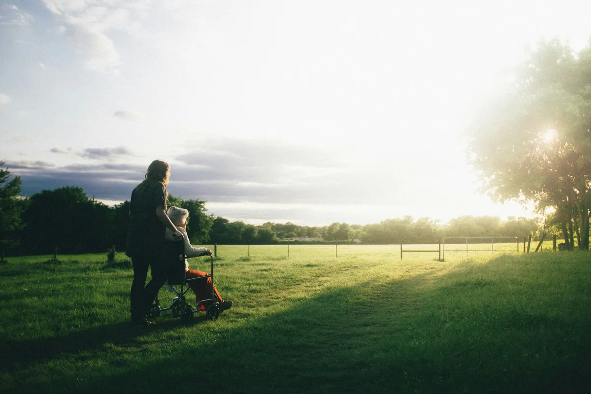 A caregiver walking a senior woman in a wheelchair across a sunlit field at golden hour