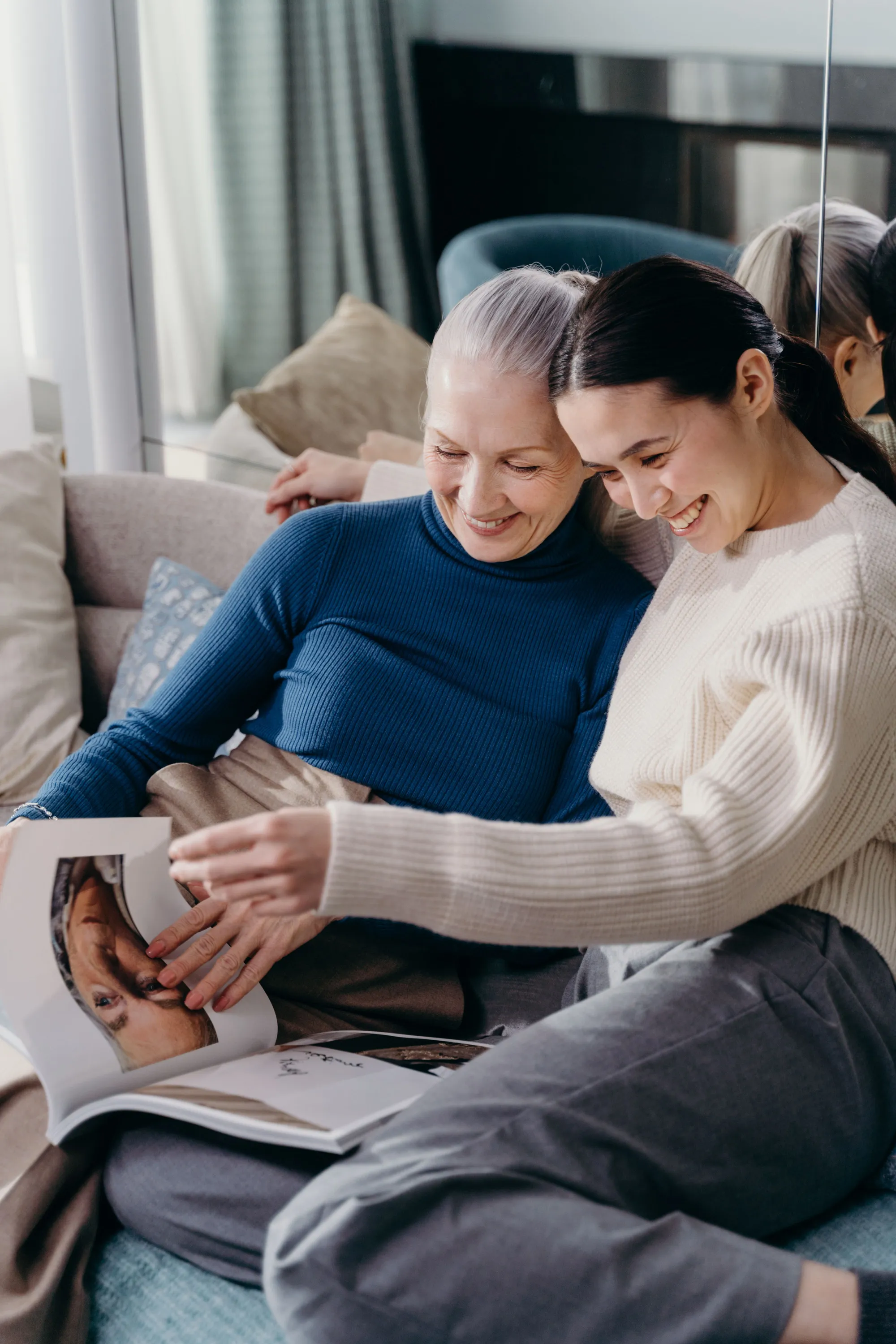 A caregiver and a senior woman laughing together over a magazine at home