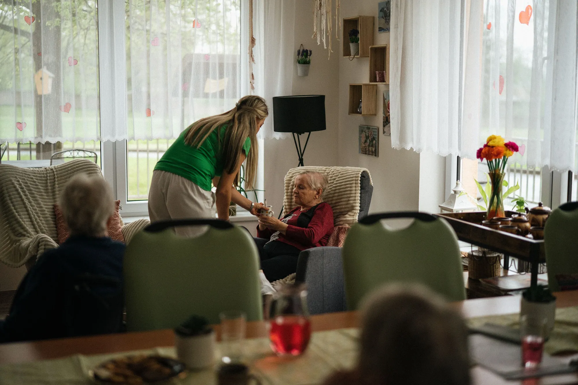 A caregiver bringing tea to a senior woman resting comfortably in an armchair