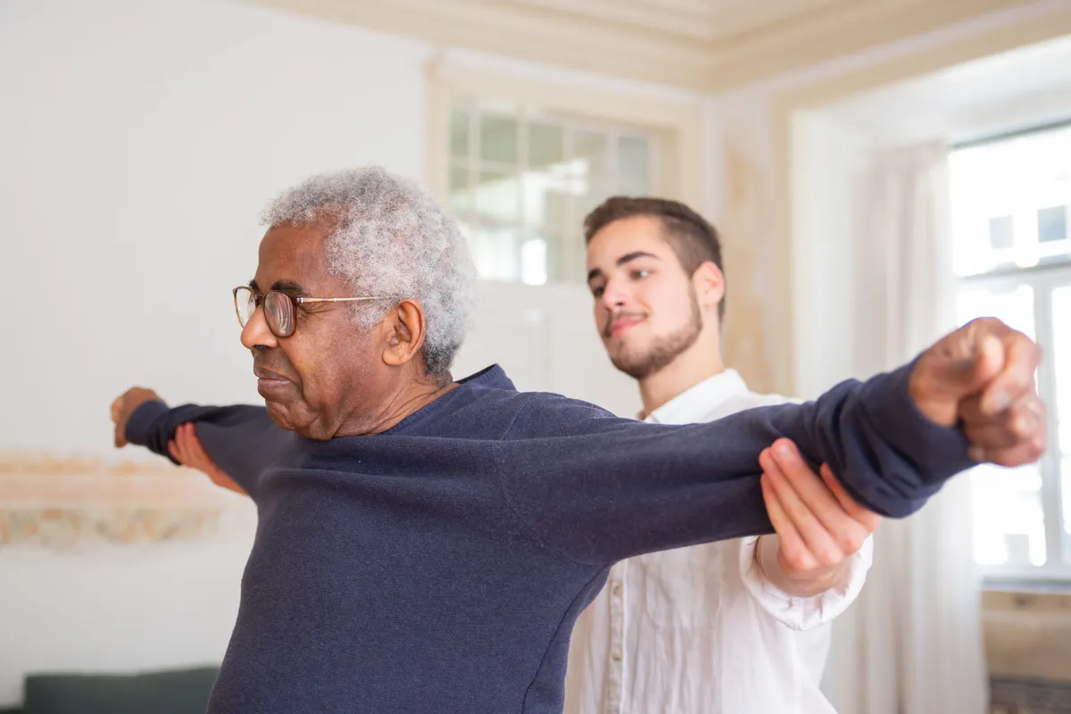 A caregiver guiding a senior man through stretching exercises in his living room