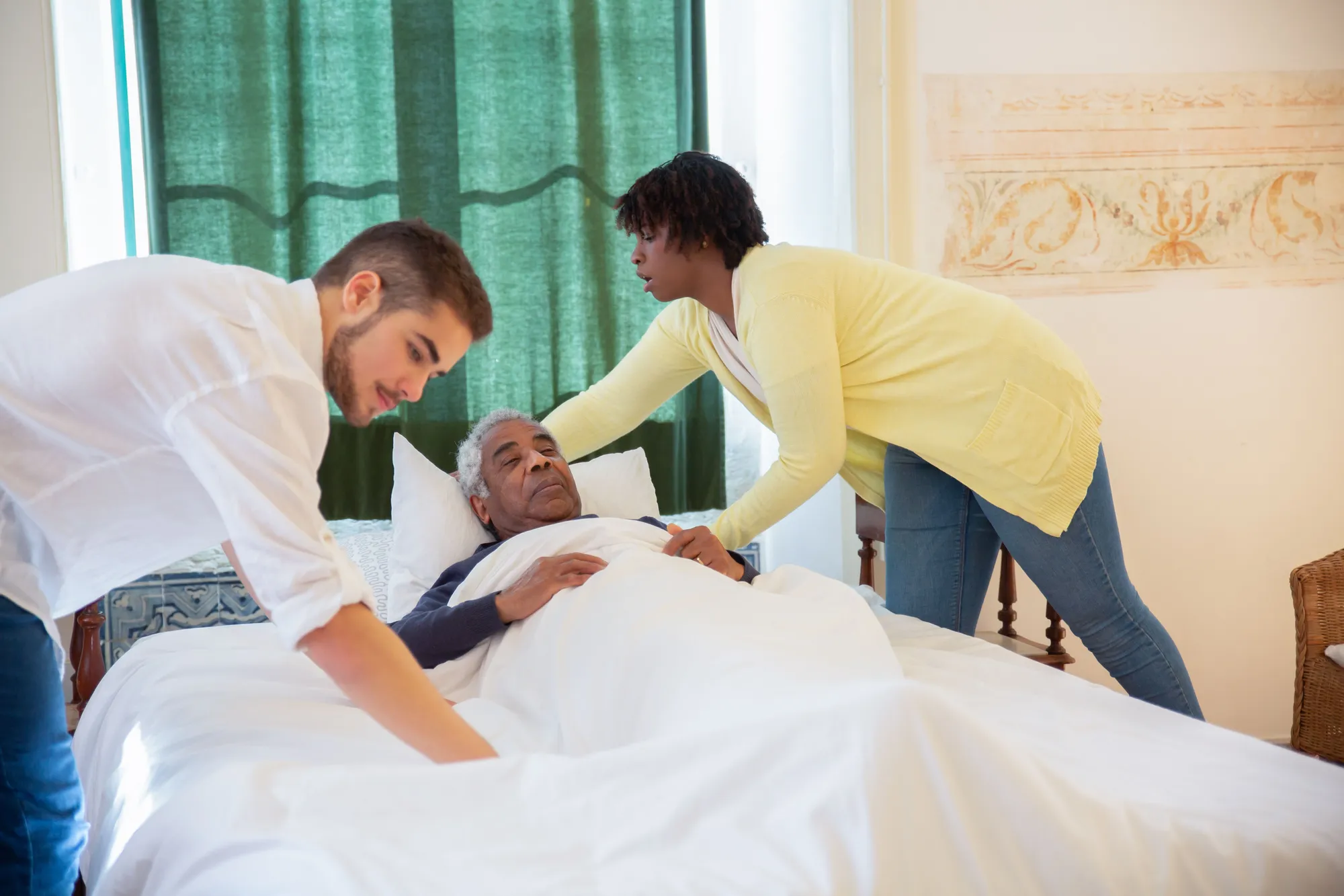 Two caregivers settling a senior man comfortably into bed