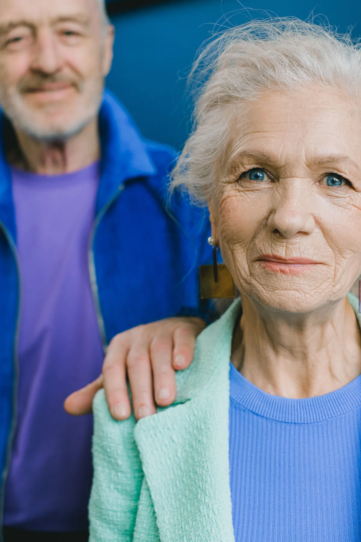 A senior woman looking ahead calmly with her husband's hand resting on her shoulder
