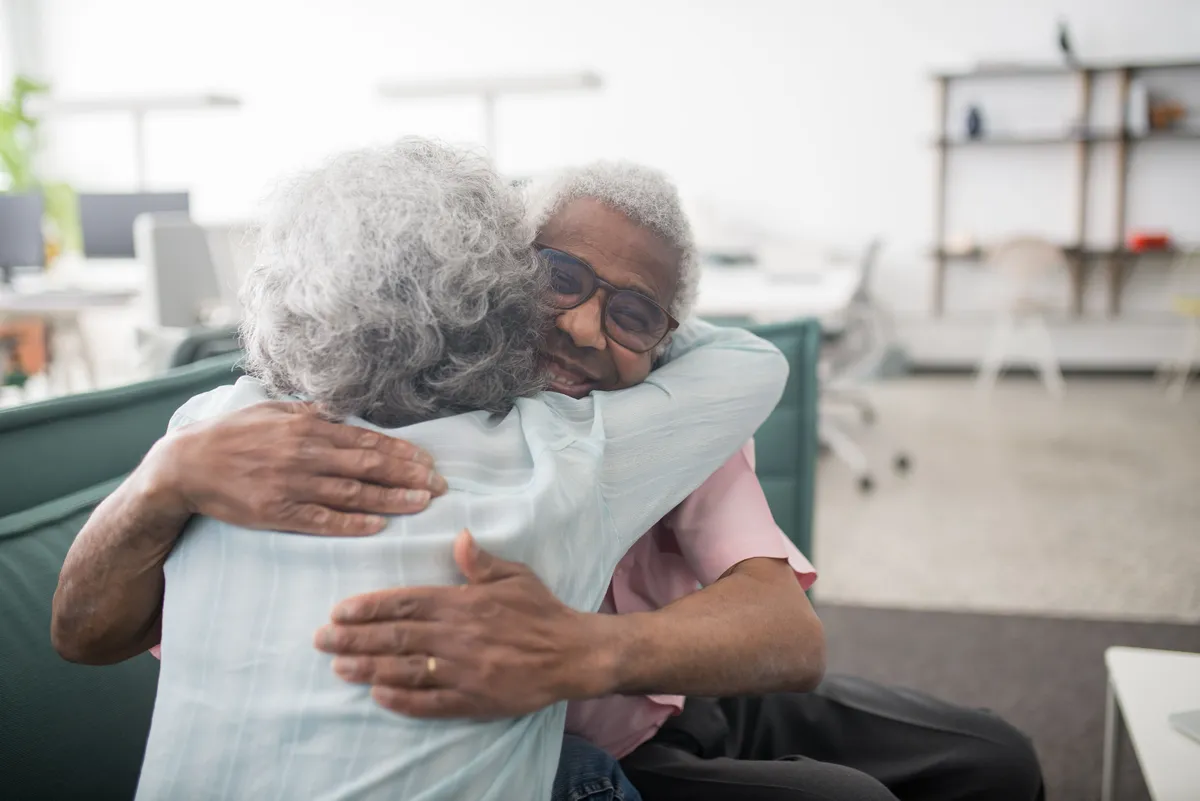 Two older adults embracing, one smiling over the other's shoulder
